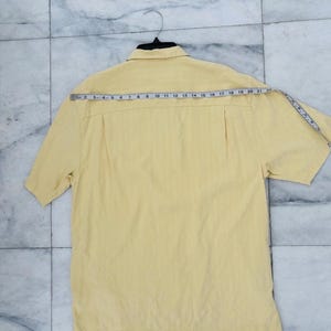 May include: A pale yellow short-sleeved shirt with a collared neckline, hanging on a black hanger. A measuring tape is draped across the back of the shirt, showing measurements in centimetres. The shirt has a vertical striped texture.
