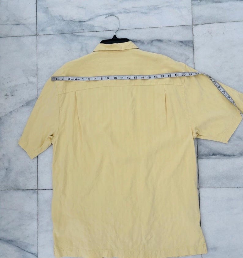May include: A pale yellow short-sleeved shirt with a collared neckline, hanging on a black hanger. A measuring tape is draped across the back of the shirt, showing measurements in centimetres. The shirt has a vertical striped texture.