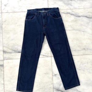 Puede incluir: Vaqueros de mezclilla azul oscuro con un diseño clásico de cinco bolsillos. Los vaqueros tienen un corte de pierna recta y cierre de botón y cremallera. La tela parece ser una mezclilla de peso medio.