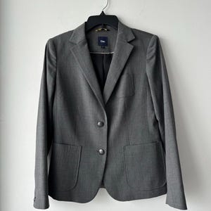 May include: A gray blazer with two buttons and two front pockets, hanging on a black hanger. The blazer has a notched lapel and a small label near the collar. The blazer is made of a textured fabric.