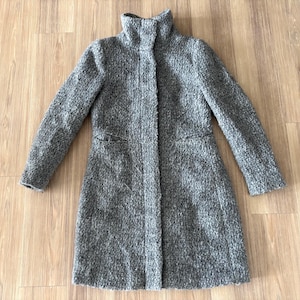 May include: A gray, textured coat with a high collar and two front pockets. The coat has long sleeves and a knee-length design. The fabric appears to be a woven material. The coat is laid flat on a wooden surface.