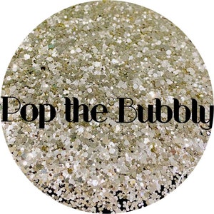 May include: A close-up of a sparkly, iridescent gold glitter. The glitter is in a round container and the text "Pop the Bubbly" is written in black letters across the glitter.