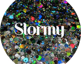 May include: A close-up of iridescent glitter in a variety of colors and shapes, including black, silver, blue, green, and red. The glitter is arranged in a circular pattern and the word "Stormy" is written in white letters in the center.