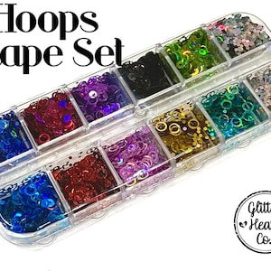 May include: A clear plastic container filled with various colors of hoop-shaped sequins. The container is divided into sections, each holding a different color, including blue, red, purple, green, and gold. The text "Hoops Shape Set" is at the top.