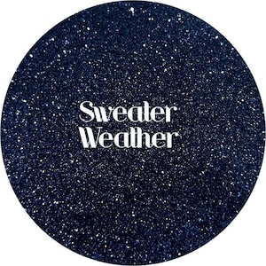 May include: A dark blue, glittery circular design with the words "Sweater Weather" in white, sans-serif font. The background is a deep navy blue with sparkling white flecks, creating a starry effect. The text is centered.