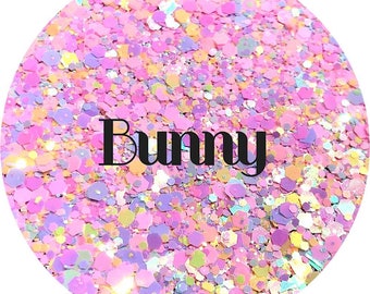May include: A circular arrangement of colorful glitter in shades of pink, purple, yellow, and white. The word "Bunny" is printed in black across the center of the glitter. The glitter pieces are various shapes and sizes.