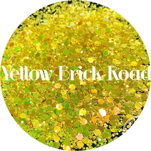 May include: Yellow glitter with a holographic finish. The glitter is in a round container and the text "Yellow Brick Road" is written in white letters across the glitter.