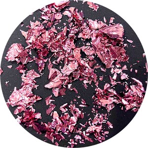 May include: Pink metallic flakes scattered on a black surface. The flakes are irregular in shape and size, creating a shimmering effect.