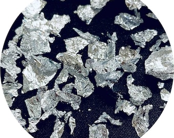 May include: Close-up of silver leaf flakes scattered on a dark surface. The flakes have a metallic sheen and irregular shapes, creating a textured appearance. The image is framed in a circular shape.