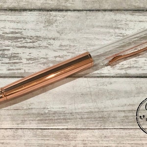 May include: A rose gold pen with a clear barrel and matching clip. The pen is lying on a light-colored, weathered wooden surface. The pen has a sleek, modern design. The pen has the words "Glitter Heart Co." in a circular logo.