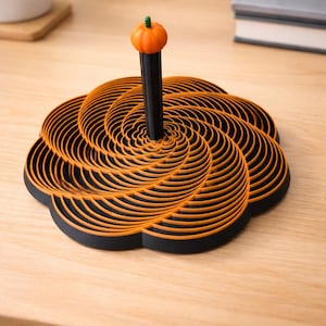 Könnte beinhalten: Ein dekorativer Halloween-Ständer mit schwarzem Sockel und orangefarbenem Spiraldesign. Ein schwarzer Pfosten erhebt sich aus der Mitte, gekrönt von einem kleinen orangefarbenen Kürbis mit grünem Stiel. Der Ständer steht auf einer Holzoberfläche.