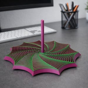 Puede incluir: Un organizador de escritorio decorativo con un diseño único en capas. La base tiene forma de estrella con capas alternas de verde y granate, enmarcadas por un borde magenta. Un poste magenta se eleva desde el centro. El organizador está sobre un escritorio gris.