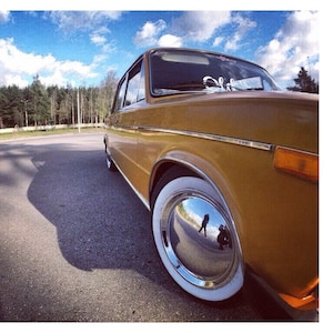 May include: A classic mustard-coloured car with a chrome hubcap and white-walled tyres. The car is parked on a paved road with a forest in the background. The sky is blue with scattered clouds.