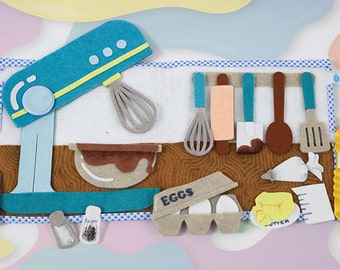 May include: A felt play set for pretend baking, featuring a blue stand mixer, a bowl of brown batter, a carton of eggs labeled "EGGS", a stick of butter labeled "BUTTER", a bottle of oil labeled "OIL", and various kitchen tools.