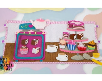 May include: A felt play set featuring a pink oven, a teapot, and a tiered cake stand with assorted cupcakes and pastries. The set is on a brown wooden surface with a pink and white checked border. The text "Titi Touch Of Art" is in the bottom left corner.