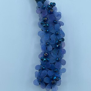 May include: A blue banana hair clip adorned with small, delicate blue fabric flowers and iridescent beads. The clip has a black base and is labeled "Blue". This hair accessory adds a touch of floral elegance.