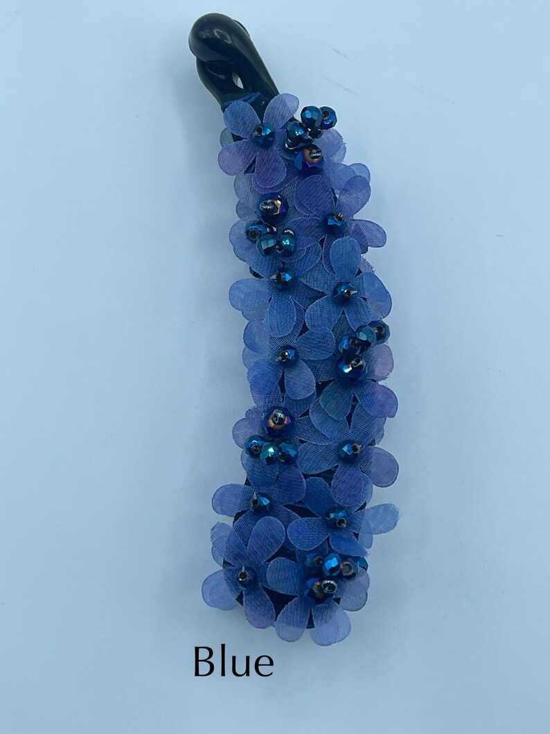 May include: A blue banana hair clip adorned with small, delicate blue fabric flowers and iridescent beads. The clip has a black base and is labeled "Blue". This hair accessory adds a touch of floral elegance.