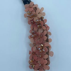 May include: A decorative hair clip with a black base, adorned with peach-colored fabric flowers and amber-colored beads. The clip is embellished with multiple layers of small, delicate flowers. The word "Orange" is printed at the bottom.