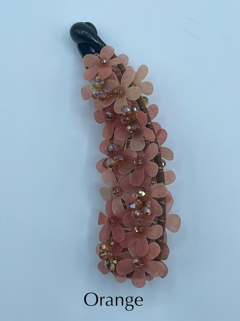 May include: A decorative hair clip with a black base, adorned with peach-colored fabric flowers and amber-colored beads. The clip is embellished with multiple layers of small, delicate flowers. The word "Orange" is printed at the bottom.