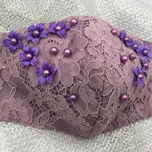 May include: A mauve lace face mask with purple flower embellishments and pearl accents.