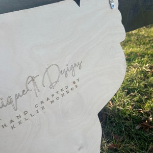 Może przedstawiać: Biały drewniany znak z wygrawerowanym tekstem "Unique Designs Hand Crafted by Kellie Monroe". Znak ma metalowy haczyk na g&oacute;rze.