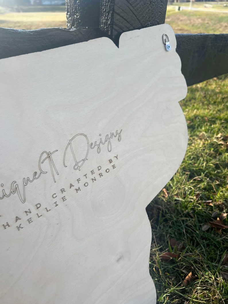 Może przedstawiać: Biały drewniany znak z wygrawerowanym tekstem "Unique Designs Hand Crafted by Kellie Monroe". Znak ma metalowy haczyk na g&oacute;rze.