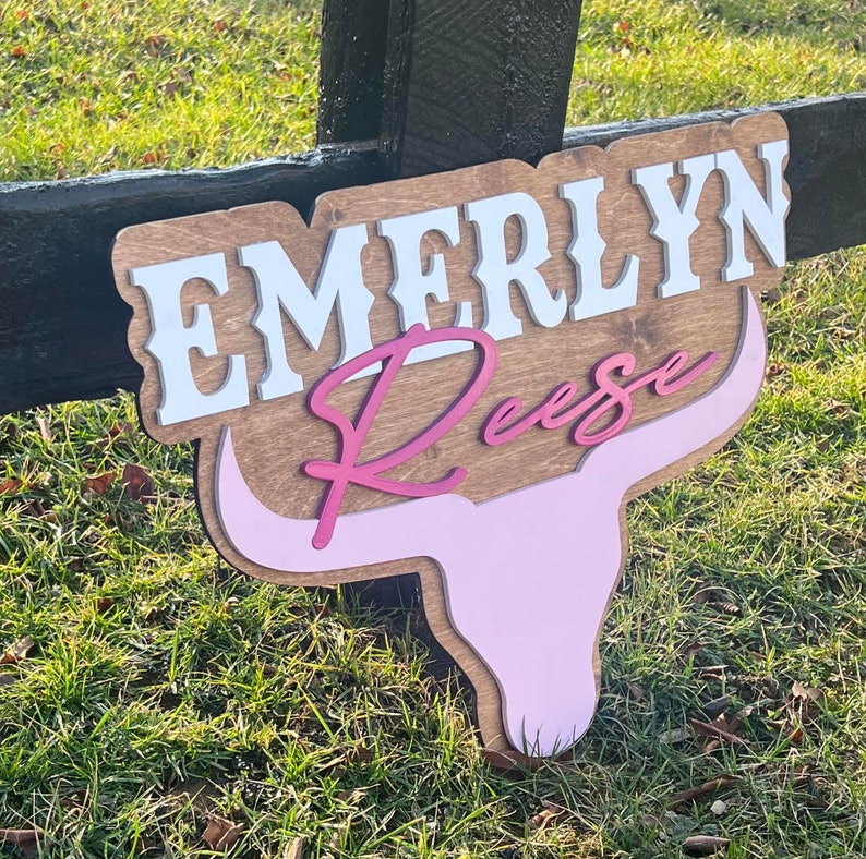 Może przedstawiać: Drewniany znak z imieniem "Emerlyn Reese" w białych i r&oacute;żowych literach. Znak ma kształt głowy krowy z r&oacute;żowymi rogami i r&oacute;żową twarzą.