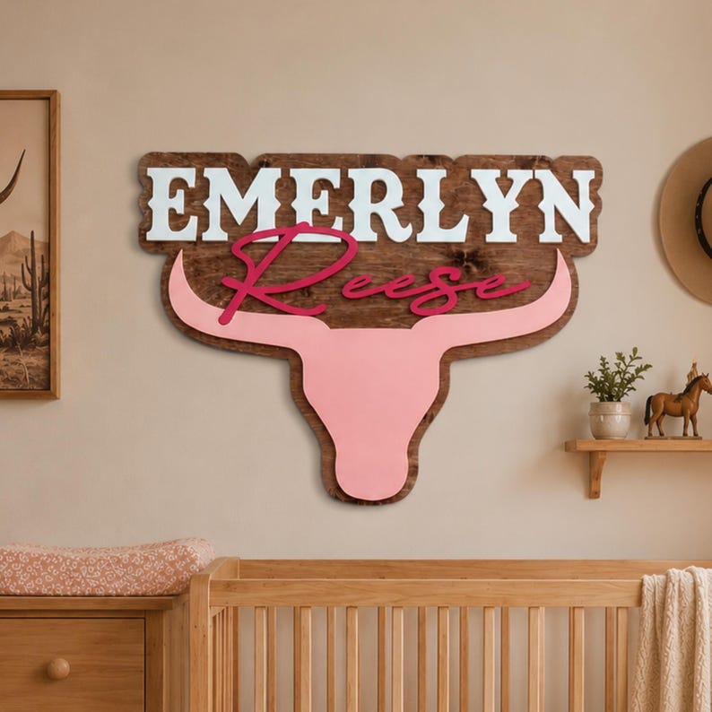 Custom western nursery name sign featuring a pink longhorn bullhead with first and middle name, outlined in wood and hanging above a crib in a neutral nursery.