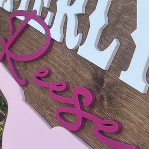 Może przedstawiać: Drewniany znak z imieniem "Emerly Reese" w białych i r&oacute;żowych literach 3D. Znak jest pomalowany brązowym wzorem drewna i ma r&oacute;żowe tło.