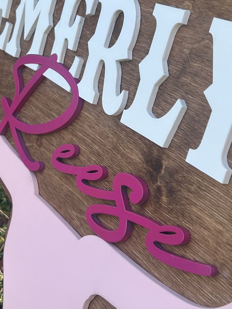 Może przedstawiać: Drewniany znak z imieniem "Emerly Reese" w białych i r&oacute;żowych literach 3D. Znak jest pomalowany brązowym wzorem drewna i ma r&oacute;żowe tło.