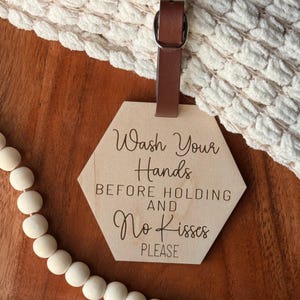 May include: A wooden, hexagon-shaped sign with the text "Wash Your Hands Before Holding and No Kisses Please." The sign is attached to a brown leather strap with a silver buckle. The background includes a white, textured blanket and wooden beads.