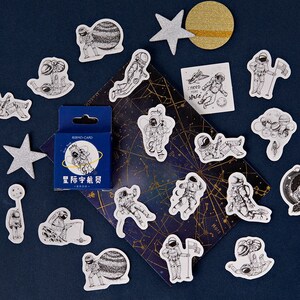 May include: A sheet of stickers featuring black and white illustrations of astronauts in various poses, including floating in space, holding a flag, and walking on a planet. The stickers are on a dark blue background with a constellation pattern. The text "I need more space" is on one of the stickers.