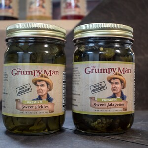 May include: Two glass jars of pickled vegetables with gold lids. The jars are labeled "The Grumpy Man" and contain "Sweet Pickles" and "Sweet Jalapeños". Both jars have a cartoon image of a man with a straw hat and the text "Made in Mississippi".