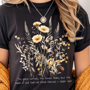 May include: A black short-sleeved t-shirt featuring a central graphic of yellow and white wildflowers and tall grasses. Below the floral design, the text reads: The grass withers, the flower fades, but the word of our God will stand forever - Isaiah 40:8.