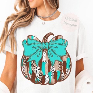May include: White t-shirt with a pumpkin design and a turquoise bow. The pumpkin has a brown stem and is decorated with leopard print, turquoise stripes, and patches of tan and white. The words "Original Designer" are printed on the shirt.