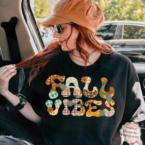 May include: Black t-shirt featuring the text "FALL VIBES" in a colourful, patterned design. The design incorporates floral and pumpkin motifs in orange, teal, and brown. The shirt is worn by a person wearing a corduroy hat and sunglasses.
