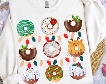 May include: A white crewneck sweatshirt featuring a graphic print of nine assorted donuts arranged in a grid. The donuts are decorated with holiday themes including icing, sprinkles, holly, a poinsettia, and a reindeer face, all connected by a string of colorful Christmas lights.