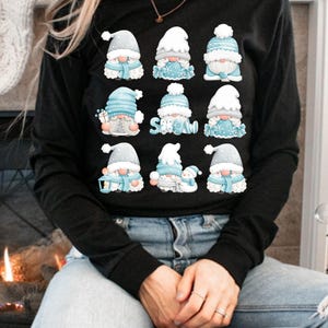 May include: A black long-sleeve shirt featuring a graphic print of nine cartoon gnomes in a grid. The gnomes wear blue and white winter hats and scarves, with some holding gifts or a snowman. The word SNOW is printed in blue letters.