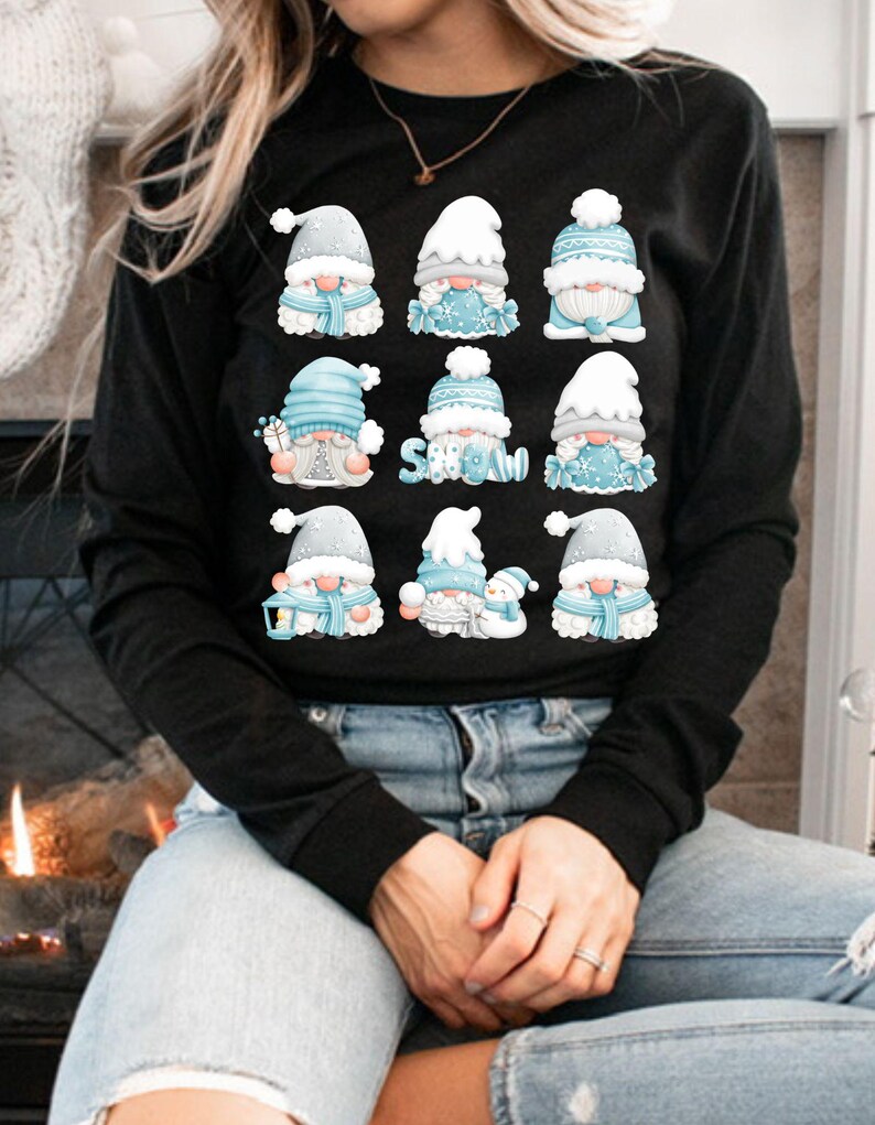 May include: A black long-sleeve shirt featuring a graphic print of nine cartoon gnomes in a grid. The gnomes wear blue and white winter hats and scarves, with some holding gifts or a snowman. The word SNOW is printed in blue letters.