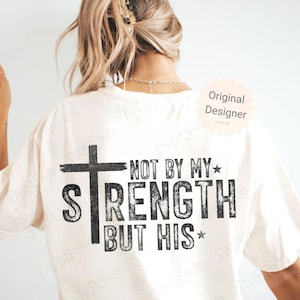 May include: Cream-colored t-shirt with a black cross and the text "NOT BY MY STRENGTH BUT HIS" in a distressed font. The shirt has a casual, relaxed fit and is suitable for everyday wear.