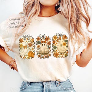 Könnte beinhalten: Cremefarbenes T-Shirt mit drei gerahmten Illustrationen. Die Illustrationen zeigen eine Gans, einen Geist und eine weitere Gans, jeweils umgeben von floralen Designs und Schleifen. Der Text "Original Design By: P31PCRAFTS" ist auf dem Shirt aufgedruckt.