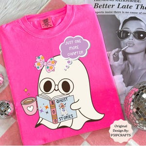 May include: A bright neon pink t-shirt featuring a graphic of a white ghost wearing glasses and a floral bow, holding a pink mug and a book titled Ghost Stories. A speech bubble above the ghost reads Just One More Chapter in white text.