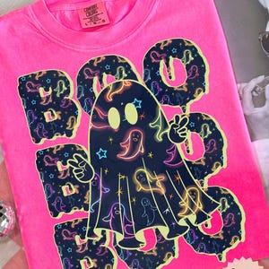 May include: A bright pink t-shirt with the word "BOO" in a spooky font, featuring a ghost design. The letters and ghost are filled with neon ghost and star graphics. The shirt has a Comfort Colors tag.