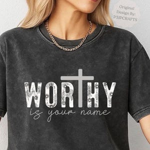 May include: A charcoal grey mineral-washed t-shirt featuring the white text WORTHY with a cross replacing the letter T. The letters are filled with a floral pattern, and the text is your name is printed in white cursive below the main graphic.