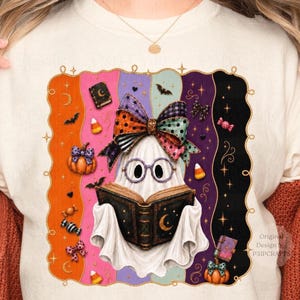 May include: A cream-coloured sweatshirt featuring a graphic of a white ghost wearing purple glasses and a large, multi-coloured patterned bow. The ghost holds an open black book with a gold moon. The background has vertical stripes and Halloween icons.
