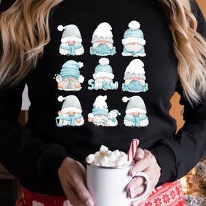 May include: A black long-sleeve shirt featuring a graphic print of nine individual gnomes in various winter outfits. The gnomes are colored in shades of light blue, white, and gray. One gnome holds a sign that says SNOW in light blue letters.