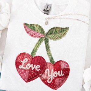 May include: A white crewneck sweatshirt featuring a large, quilted cherry design on the front. The two cherries are shaped like hearts, colored in various shades of red and pink, with the words Love and You embroidered in white script.