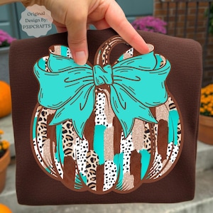 May include: A dark brown t-shirt featuring a graphic of a pumpkin decorated with a large turquoise bow. The pumpkin design incorporates vertical stripes of leopard print, white with black spots, and glittery tan textures on a dark brown background.