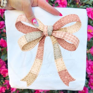 May include: A fabric bow featuring a patchwork design with alternating sections of muted pink, beige, and sage green fabric. The bow is constructed with visible stitching along the edges and includes two long, dangling ribbon tails with notched ends.