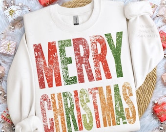 May include: A white long-sleeve crewneck sweatshirt featuring the text MERRY CHRISTMAS printed in a distressed, multi-colored font. The letters alternate between shades of red, green, and gold, creating a textured, weathered appearance.